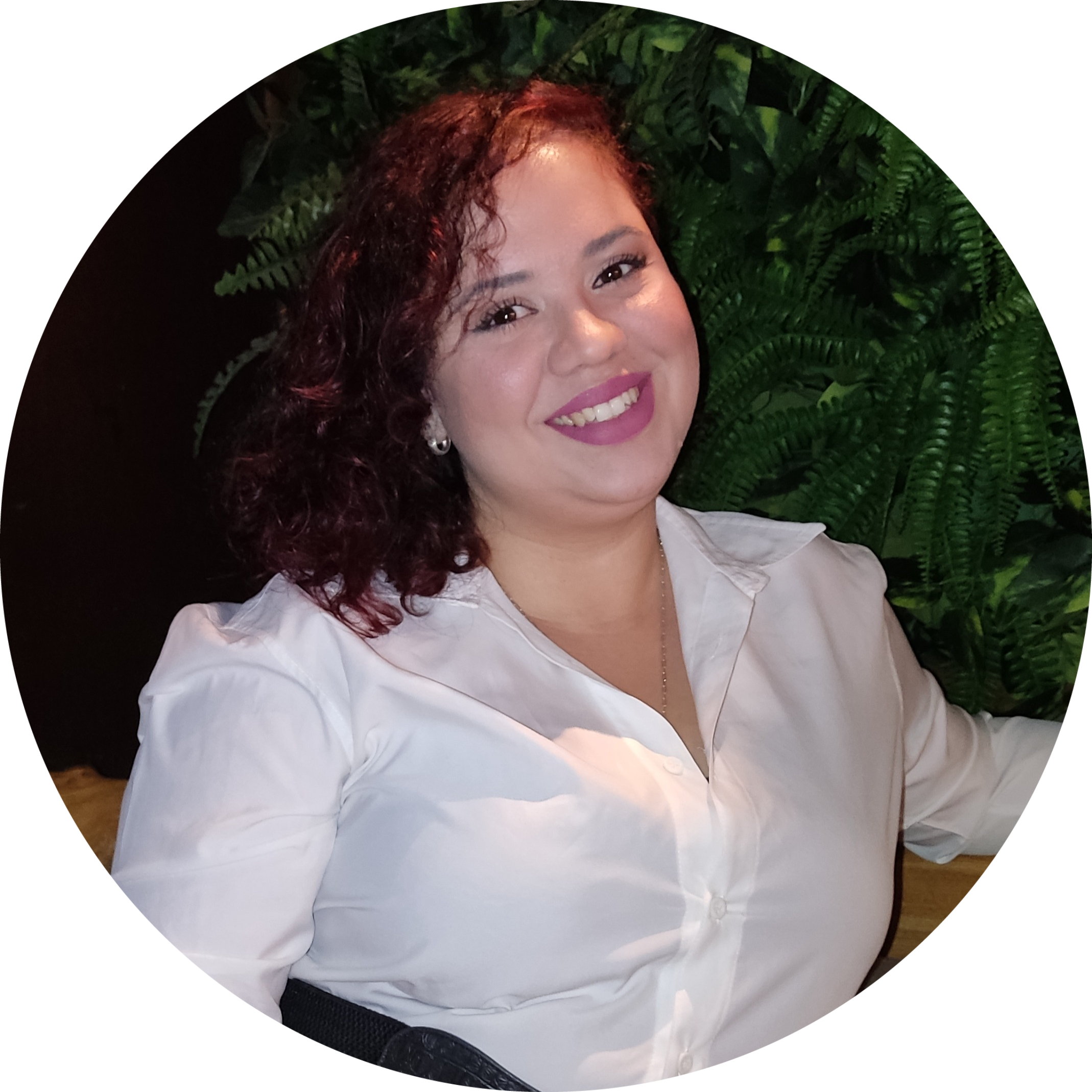 Mulher branca de cabelos vermelhos, com camisa branca sentada com uma parede de fundo coberta de plantas